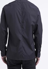 Chemise noire à manches longues avec une coupe droite, des poignets boutonnés, et une texture lisse et mate. Dispose d'une couture centrale le long du dos.