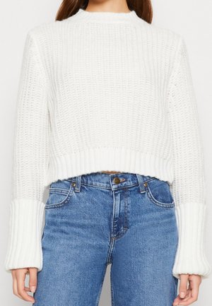Pull en maille épaisse blanche avec larges poignets côtelés, associé à un jean bleu taille haute, coupé à la taille.