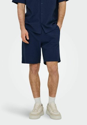 Homme portant un short bleu marine et une chemise à manches courtes boutonnée assortie, avec des chaussettes blanches et des baskets beige clair.