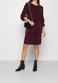 Mauve Faltenkleid mit einem Boot-Ausschnitt, drei Viertel-Ärmeln und geschichteten Design. Kombiniert mit schwarzen Knöchelboots und einer strukturierten Handtasche.