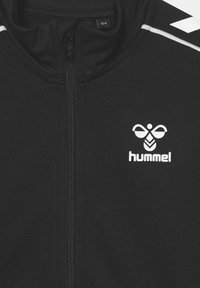 Chaqueta negra con cremallera completa, que presenta el logo de Hummel en blanco y franjas de acento en los hombros. Textura de tela suave, ajuste atlético.