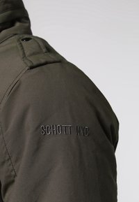 Épaule de veste vert foncé avec texte noir cousu "SCHOTT NYC" et un détail de sangle en tissu boutonné près du col.