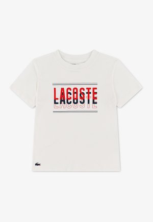 T-shirt blanc à manches courtes avec un texte superposé rouge et bleu marine "LACOSTE" et un petit logo crocodile sur l'ourlet inférieur gauche.