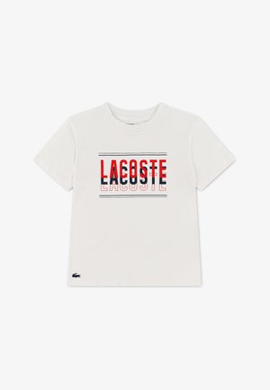 T-shirt blanc à manches courtes avec un texte superposé rouge et bleu marine "LACOSTE" et un petit logo crocodile sur l'ourlet inférieur gauche.