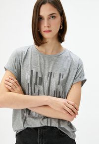 Grå kortærmet t-shirt med et vertikalt stribet sølv grafikdesign. Blødt stof, afslappet pasform, rund halsudskæring.