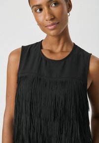 Ženska z obročnimi uhani, oblečena v brezrokavnico črne barve z slojnimi frinžami, ki rahlo gleda na stran pred enobarvno ozadje.