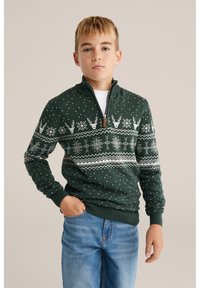 Mörkgrön stickad tröja med vita mönster av snöflingor och renar, med dragkedja vid kragen och ribbade muddar. Kombinerad med blå jeans.