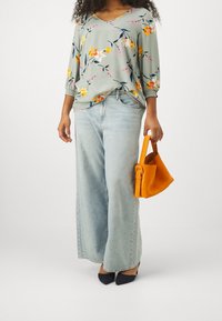Blusa con stampa floreale in verde chiaro con maniche a tre quarti, abbinata a pantaloni in denim a gamba larga blu chiaro e una borsa in suede arancione. Tacchi neri.