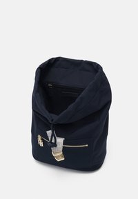 Mochila de tela azul marino con cierre de cordón, bolsillo frontal con cremallera, herrajes dorados y textura suave. Diseño práctico y compacto.
