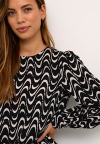 Schwarzes Kleid mit weißem Wellenmuster, langen Puffärmeln, runder Ausschnitt und weicher Textur. Das Model posiert mit einer Hand in die Hüfte gestützt.