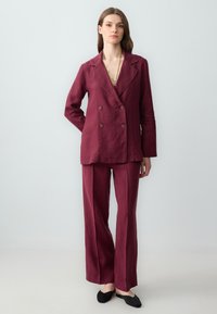 Burgundowa lniana marynarka z podwójnymi guzikami, dopasowane szerokie spodnie oraz czarne buty wsuwane. Prosta złota naszyjnik jako akcent.