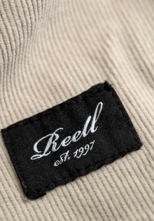 Black fabric tag with white embroidery reading "Reell est. 1997" on a textured beige corduroy background.