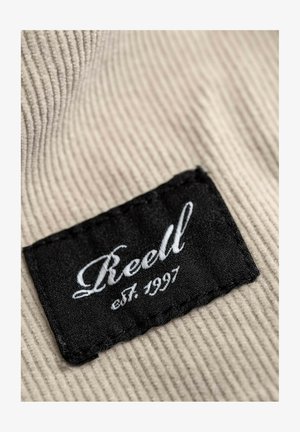 Schwarzes Stoffetikett mit weißer Stickerei, auf der "Reell est. 1997" steht, vor einem strukturierten beigefarbenen Cordstoff-Hintergrund.