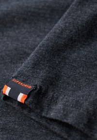 Acercamiento de una tela gris oscuro con un borde doblado y una etiqueta negra con el texto naranja "SUPERDRY®" y rayas naranjas y blancas.
