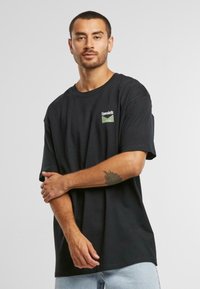 Black cotton t-shirt with short sleeves, featuring a green logo patch on the chest, round neckline, and relaxed fit.