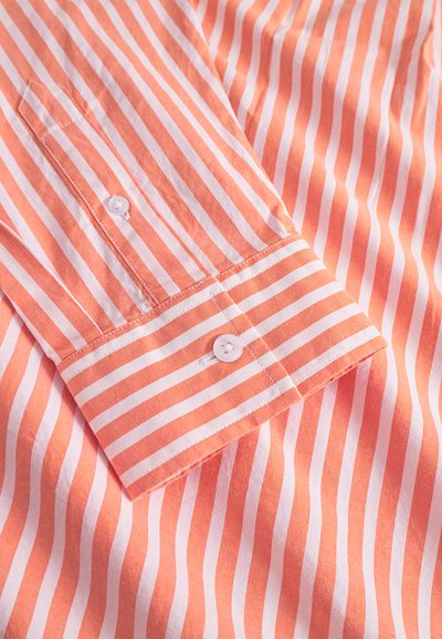 Close-up of a orange and white vertically striped shirt sleeve with cuff and white button, showing fabric texture and stitching details.