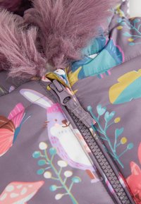 Nærbillede af en farverig børnejakke lynlås med en kanin- og naturprint, med to lilla fluffy pomponer fastgjort til lynlåstrækkeren.