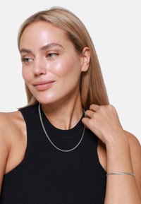 Collana di catena argento e braccialetto abbinato indossati da una persona, con una canotta nera a coste senza maniche e lunghi capelli biondo chiaro.