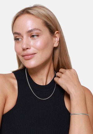 Collana di catena argento e braccialetto abbinato indossati da una persona, con una canotta nera a coste senza maniche e lunghi capelli biondo chiaro.