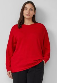 Rode geribbelde longsleeve top met een ronde hals en een losse pasvorm, gecombineerd met donkere broeken. De stof heeft een subtiele textuur en valt elegant.