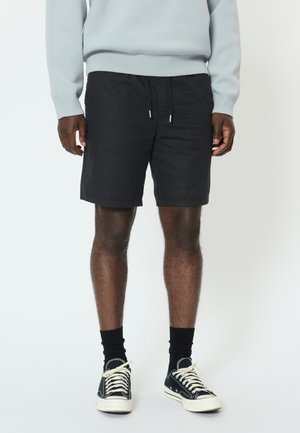 Bărbat purtând pantaloni scurți negri cu șnur, șosete negre până la gleznă, adidași negri și albi și un hanorac gri deschis, stând pe un fundal alb.