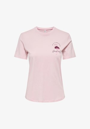 Giovane donna sorridente, che indossa una t-shirt grafica rosa chiaro, jeans neri e stivali neri a punta, in piedi con le braccia incrociate.