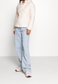 Světle béžová zateplená bunda s kapucí, prošívaný design, plný zip, nošená přes fialový top, v kombinaci se světle modrými širokými džínami a teniskami.