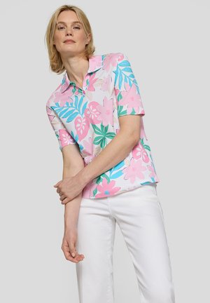 Frau trägt ein kurzärmeliges Hemd mit pink-blauem Blumenmuster und weiße Hosen und steht vor einem einfarbigen hellgrauen Hintergrund.