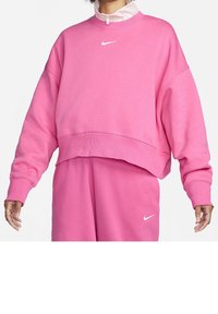 Osoba oblečená v jasně růžové mikině Nike a odpovídajících teplácích, obě s bílými logy swoosh, ruce viditelné po stranách.