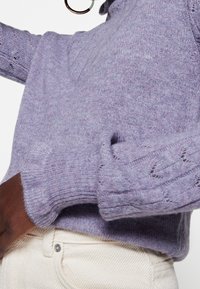 Lavendelfärgad stickad tröja med mönstrade ärmar och ribbade muddar. Mjuk textur och figurnära design, kombinerad med ljusa jeans.