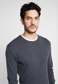 Dunkelgraues Langarmshirt mit rundem Halsausschnitt und weißen Akzenten. Glatte Stricktextur, taillierter Schnitt betont den Oberkörper.