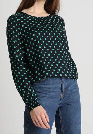 Vrouw die een zwarte blouse met lange mouwen met groene stippen draagt en blauwe high-waisted jeans, staand tegen een effen achtergrond.