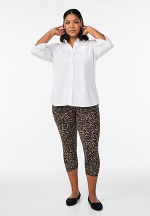 Žena podešava kosu, nosi bijelu košulju s gumbima, kratke helanke s uzorkom leoparda i crne baletanke na jednobojnoj pozadini.