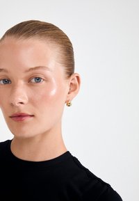 Χρυσά σκουλαρίκια σε σχήμα σταγόνας αντιτίθενται σε ένα μαύρο τοπ. Το δέρμα είναι λείο με μια διακριτική λάμψη; τα μαλλιά είναι τραβηγμένα προς τα πίσω για ένα καθαρό look.