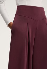 Pantaloni largi burgundy, confecționați dintr-un material moale, au un talie înaltă, buzunare laterale adânci și plii subtile pentru un plus de structură.