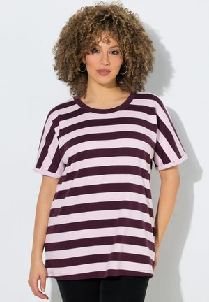 Vrouw met krullend haar die creolen draagt en een shirt met korte mouwen met roze en donkerpaarse horizontale strepen, staand tegen een witte muur.