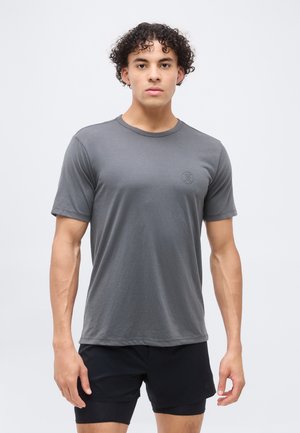 Młody mężczyzna z kręconymi włosami w prostym szarym t-shircie i czarnych szortach, stojący na jednolitym jasnym tle.