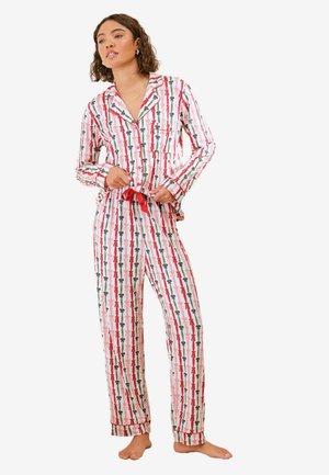 Woman wearing white pajamas with vertical red and green bow patterns, adjusting the red drawstring on the pajama pants.