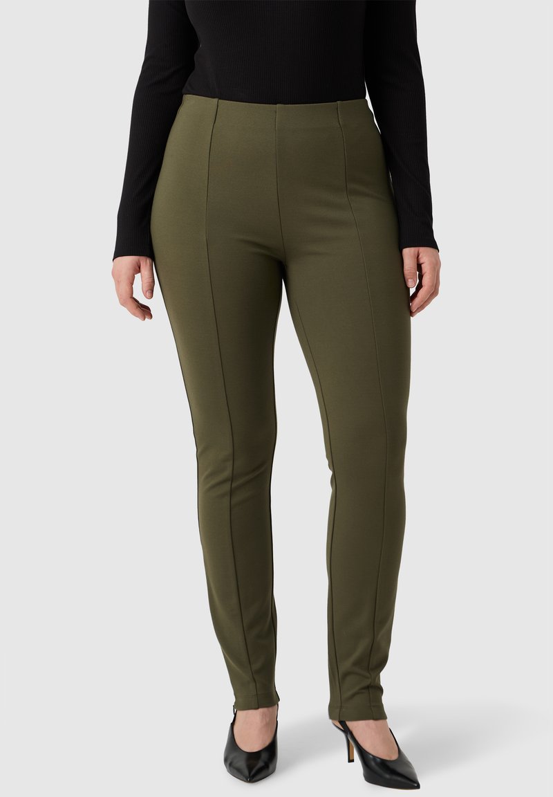 Donna che indossa pantaloni aderenti verde oliva con cuciture frontali, una maglietta nera a maniche lunghe e tacchi alti neri a punta, in piedi su uno sfondo grigio.