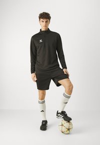 Cămașă sport neagră cu mânecă lungă și fermoar, pantaloni scurți negri, șosete albe cu dungi negre și ghete negre; stând pe o minge de fotbal.