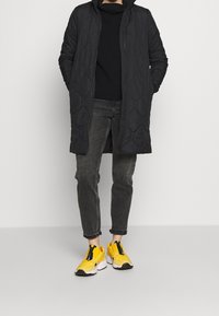 Czarny pikowany długi płaszcz, czarny sweter z dzianiny, szare dżinsy oraz jasnoszare klapki w żółtym kolorze z czarno-białymi podeszwami.