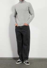 Homme portant un pull col roulé gris clair, un jean noir ample et des baskets lacées noir et blanc, se tenant avec les mains dans les poches.