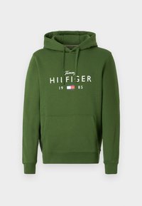 Haut à capuche vert avec une poche kangourou, capuche ajustable avec cordon de serrage, et texte imprimé en blanc "Tommy HILFIGER 1985" avec un logo de drapeau.