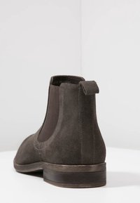 Botín Chelsea de ante marrón oscuro con panel elástico lateral y tirador, visto desde atrás sobre una superficie blanca contra un fondo liso.