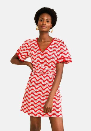 Nő piros-fehér kagylómintás rövid ruhát visel, pöttyös ujjakkal és karika fülbevalóval, egyik kezét a csípőjén tartva áll.