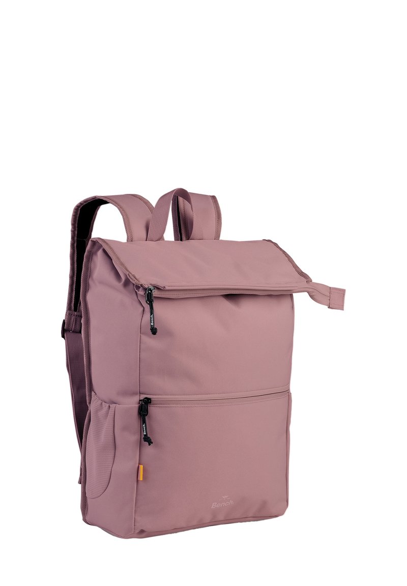 Sac à dos en tissu rose avec une fermeture roulée, des bretelles ajustables, deux poches zippées à l'avant et une poche en mesh sur le côté. Texture lisse, design minimaliste.