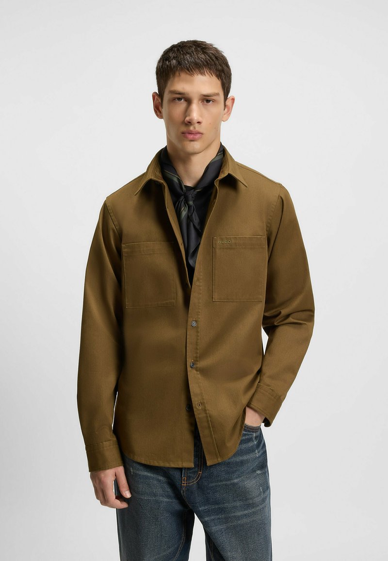 Brun, langærmet skjorte med en glat tekstur og to brystlommer. Båret over en mørk skjorte, parret med blå jeans. Detaljer med sort tørklæde.
