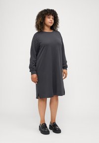 Grijs langemouwen jurk met studs op de schouders, zijsplits aan de zoom, gemaakt van zachte stof, gedragen met zwarte schoenen met studs.