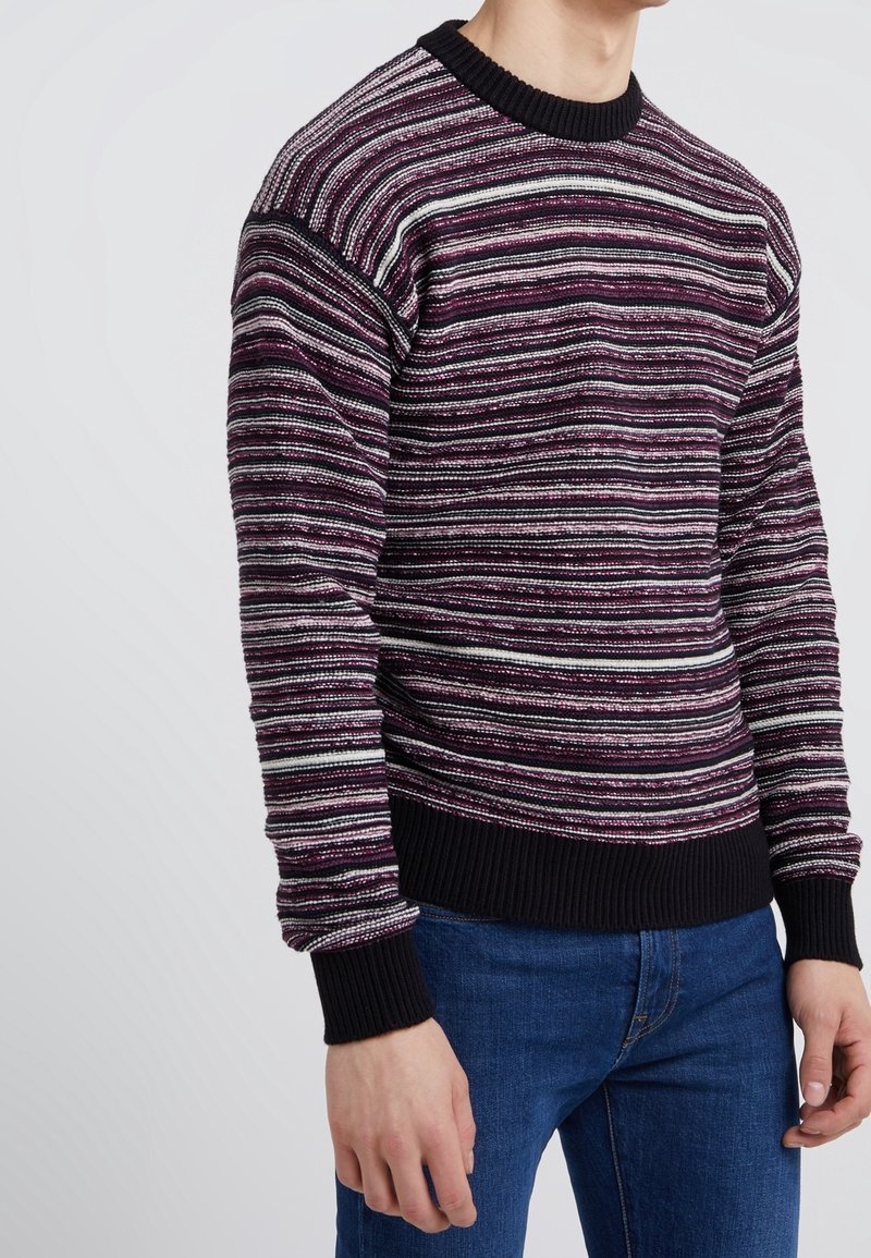 Man draagt een gebreide trui met horizontale strepen in donkerpaars, zwart en wit, met zwarte geribbelde manchetten en zoom, gecombineerd met een blauwe spijkerbroek.