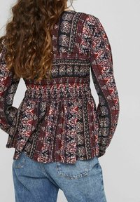 Bloes met bloemenpatroon in diepe roodtinten, zwart en wit, met smockdetails in de taille, lange mouwen en een peplum-silhouet.